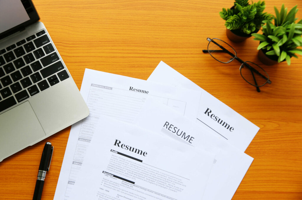 Resumes on a table with laptop, glasses and pen.